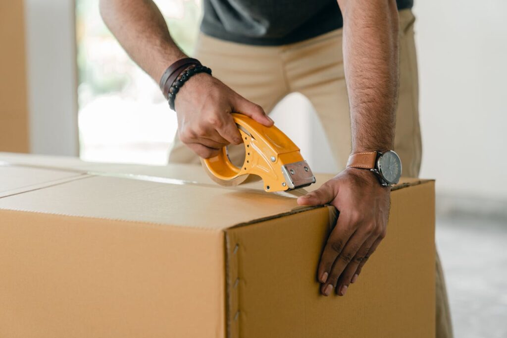 Evitar el estrés a tu mascota durante la mudanza Crop faceless young male with wristwatch using adhesive tape while preparing cardboard box for transportation